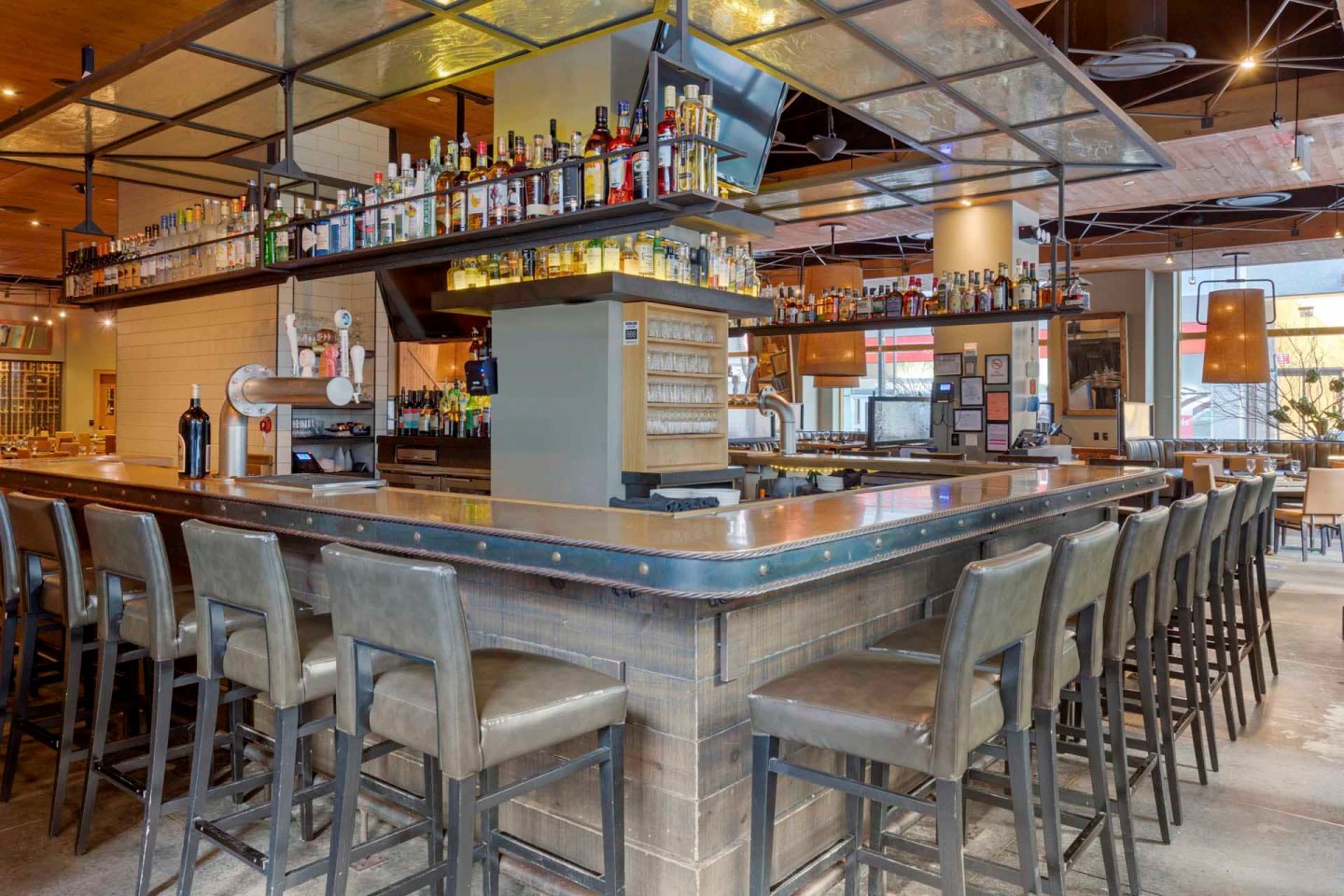 A modern bar with a U-shaped counter surrounded by high chairs. The shelves above are stocked with various bottles. The interior features industrial-style decor with metal accents and warm lighting. Tables and seating are visible in the background.