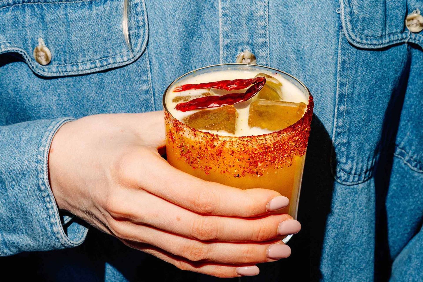 A hand with light pink nails holds a glass of orange cocktail with ice, a chili powder rim, and garnished with dried chili peppers. The person is wearing a blue denim shirt.