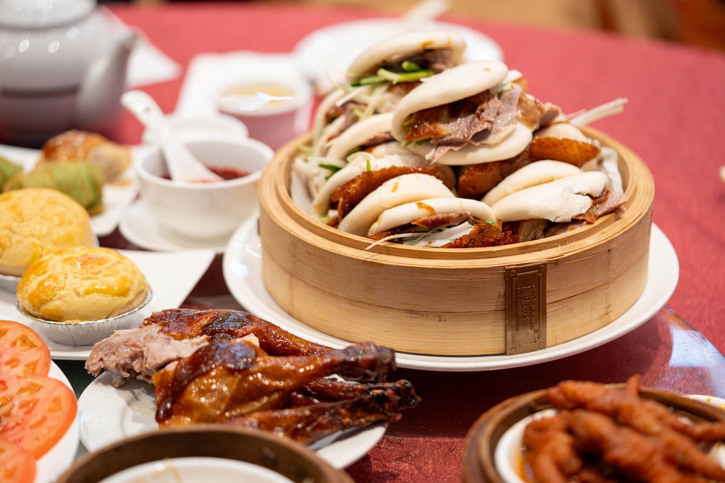 A bamboo steamer filled with stacked bao buns sits on a table with roasted duck, dim sum, sauces, tomatoes, and pastries, ready to be enjoyed at a Chinese meal.