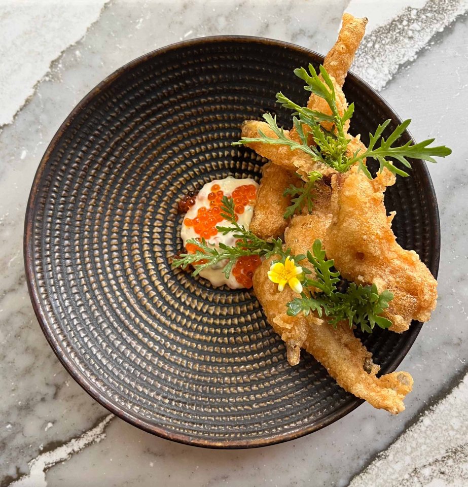 A patterned bowl with fried frog legs garnished with greens and a yellow flower, served on a dollop of creamy sauce topped with orange fish roe, on a marble surface.