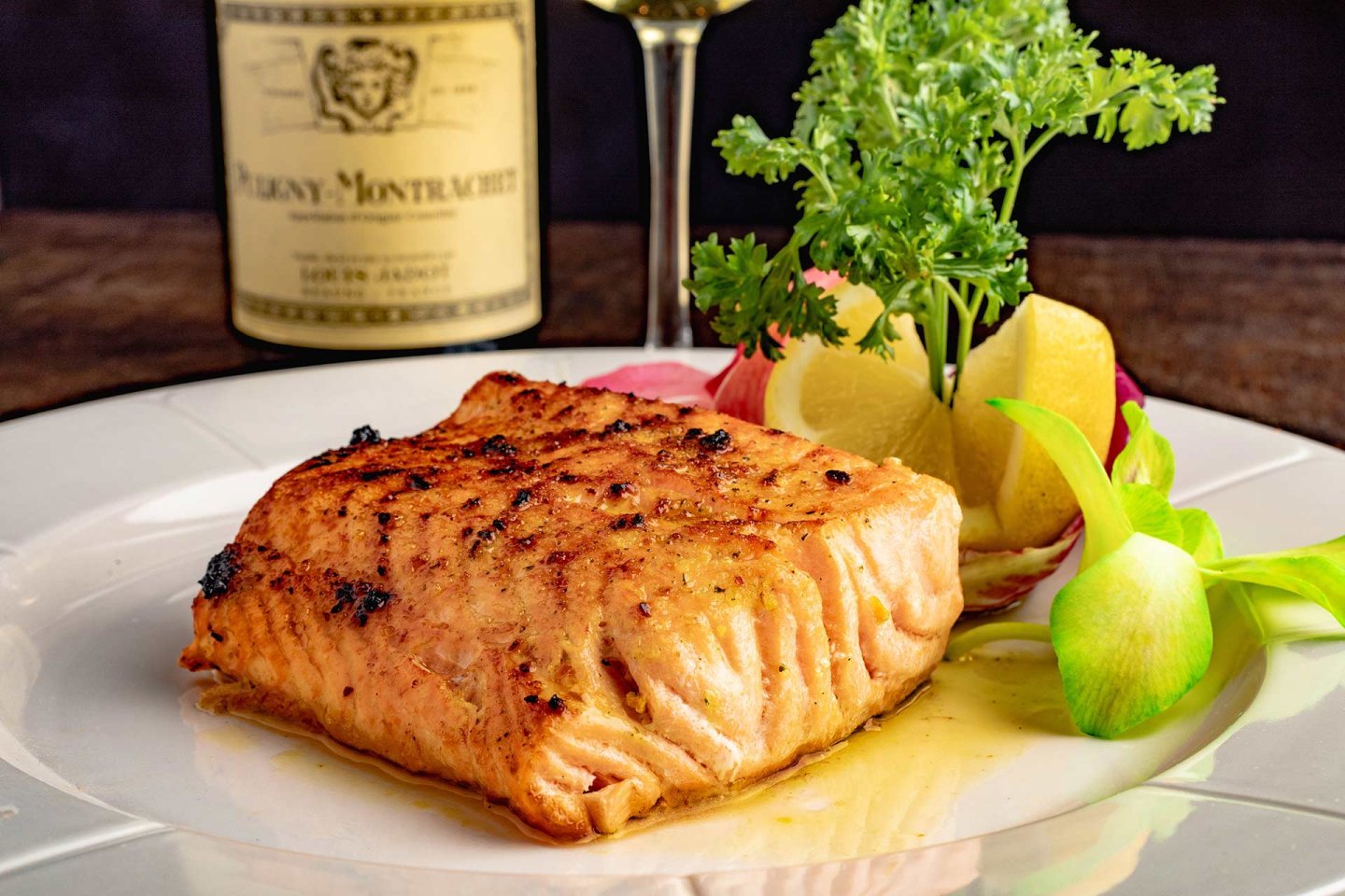 Grilled salmon fillet on a white plate, garnished with parsley, lemon wedges, and vegetables. A bottle of white wine and a wine glass are blurred in the background.