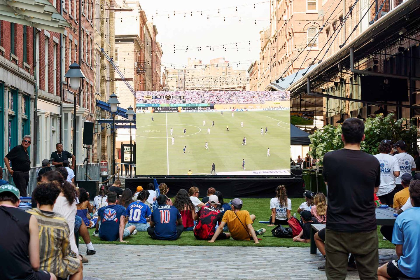 A crowd sits outdoors on a cobblestone street watching a large screen showing a soccer match, surrounded by brick buildings and string lights hanging overhead.