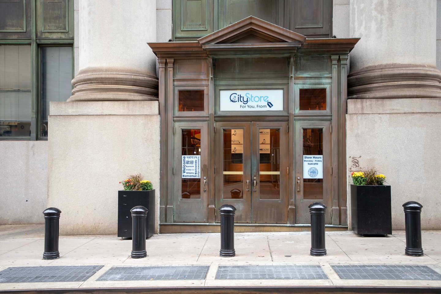 Entrance to a CityStore with glass doors, signs, and flower planters on each side, set in a tall stone building with columns. Five black bollards line the sidewalk in front.