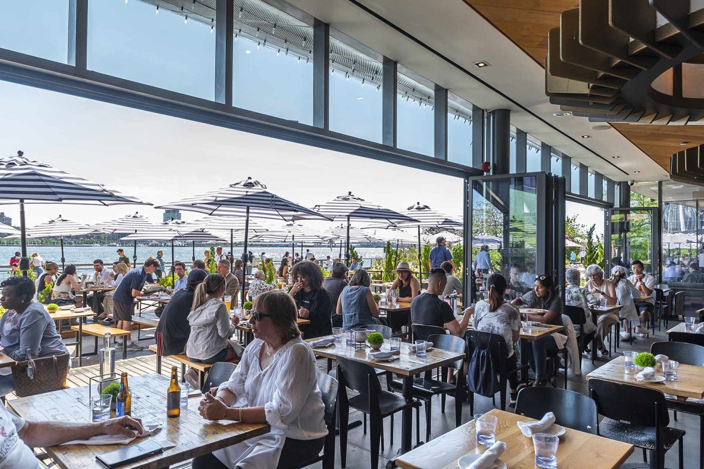 People dine at a busy, modern restaurant with large windows and outdoor seating. Black-and-white striped umbrellas shade the patio; sunlight streams in, and greenery surrounds the waterfront view.