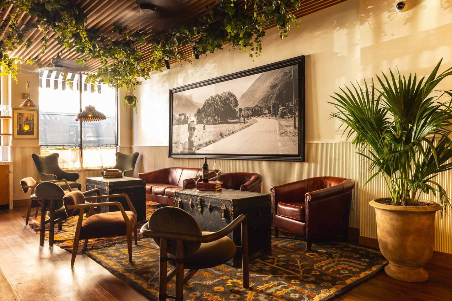 A cozy lounge with leather chairs, a sofa, a large trunk as a coffee table, potted plants, hanging greenery, and a framed black-and-white landscape photo on the wall, with warm sunlight streaming through the window.