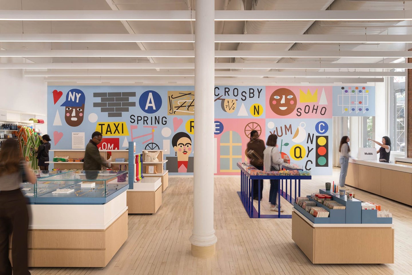 People browse a brightly lit store with high ceilings, wooden floors, and colorful, geometric wall art featuring faces, letters, and symbols. Display cases and tables hold various items for sale.