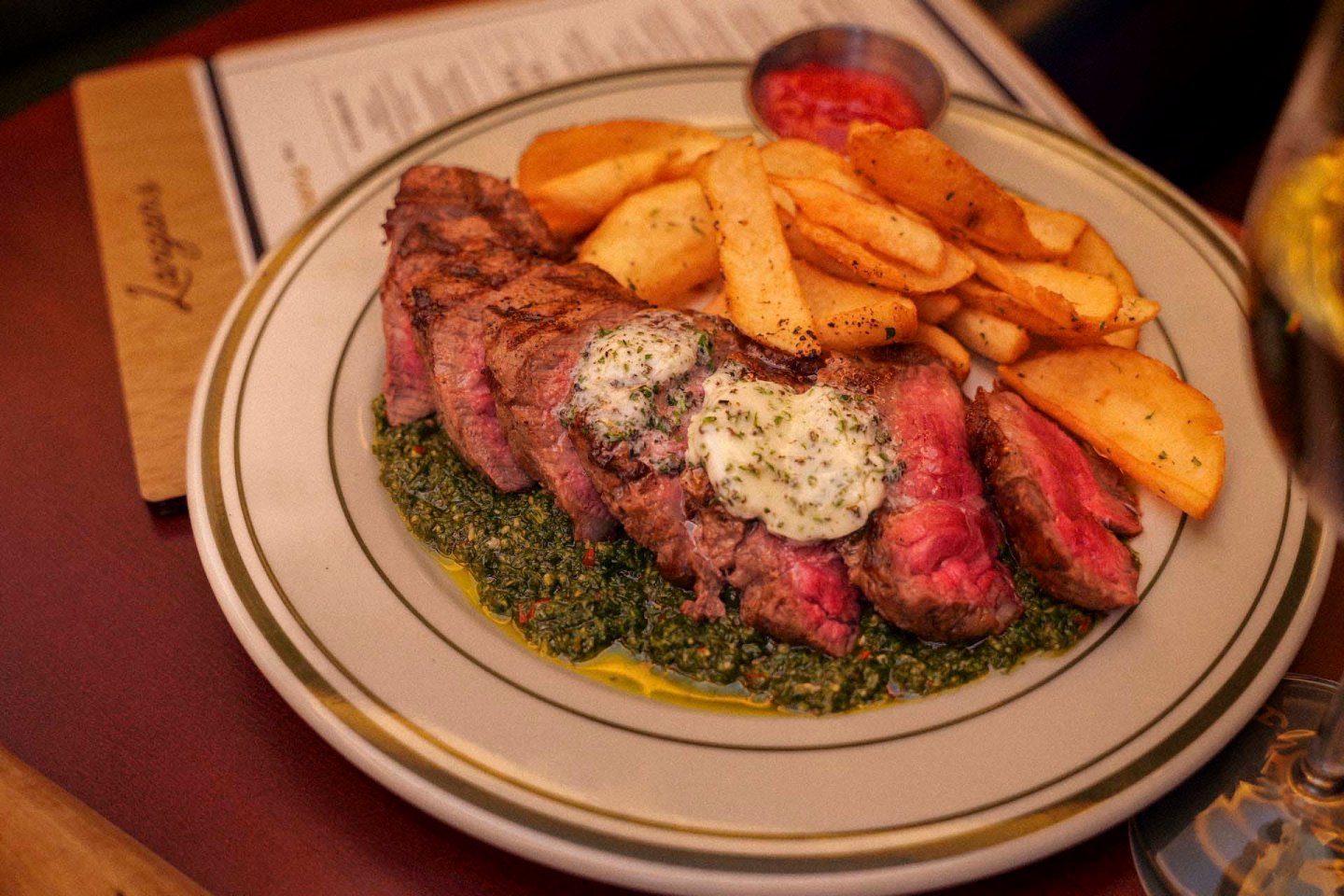 A plate of sliced medium-rare steak topped with herbed butter, served with thick-cut fries and a green pesto sauce. A small dish with red sauce is in the background, with a menu partially visible on the table.