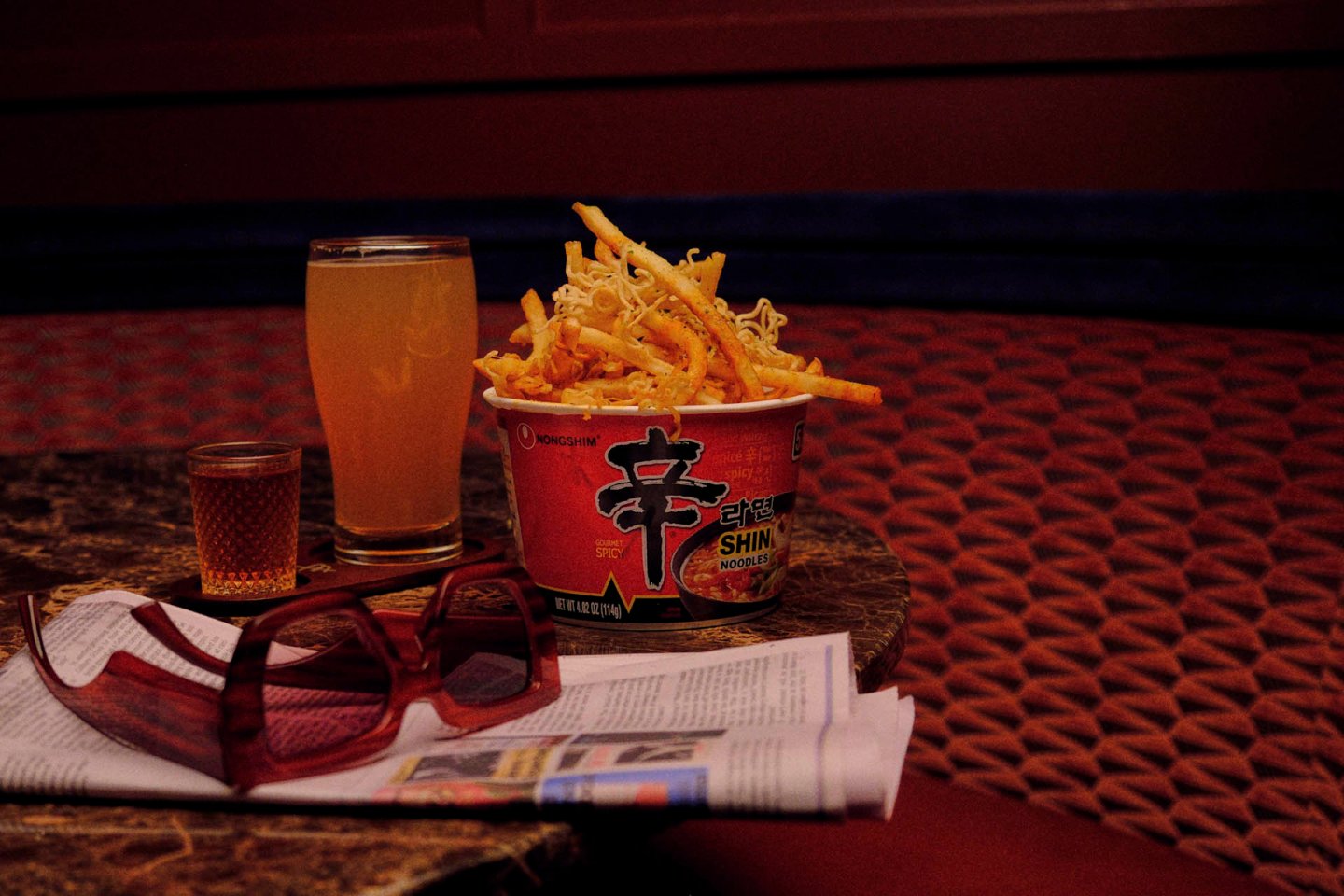 A cup of spicy Shin Ramyun with crispy toppings and two glasses of tea placed on a table. A folded newspaper and sunglasses are nearby. The setting has a warm, cozy ambiance with patterned seating in the background.