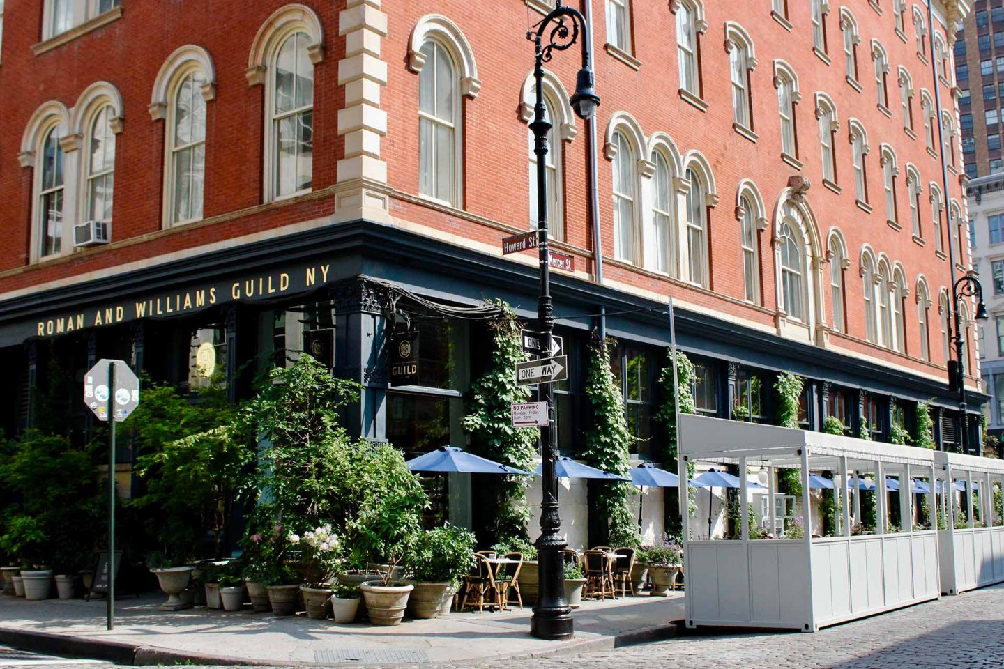 Street view of a red-brick corner building with arched windows, a black storefront labeled "Roman and Williams Guild NY," outdoor dining area with blue umbrellas, greenery, and street signs on a sunny day.