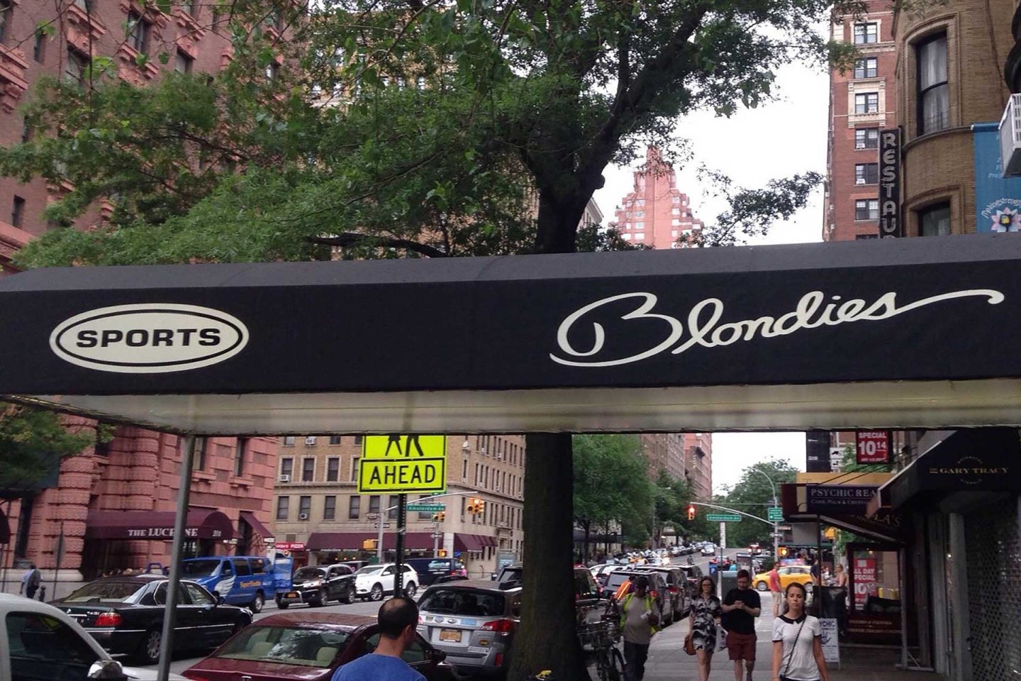 A black awning with "SPORTS Blondies" written in white hangs over a busy city sidewalk with pedestrians, parked cars, trees, and tall brick buildings in the background.