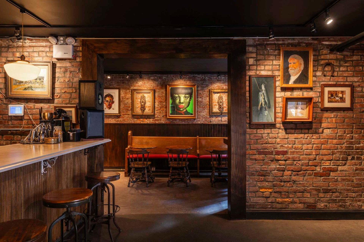 A cozy, dimly lit bar with exposed brick walls. Wooden stools line the counter on the left, while the seating area in the back features a wooden bench and chairs. Various framed portraits and artwork adorn the walls.