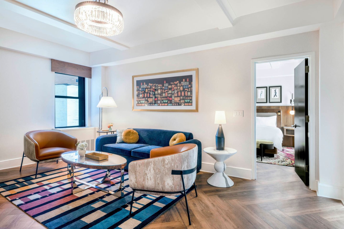 Modern hotel suite living area with a blue velvet sofa, two brown chairs, a round coffee table, a colorful rug, a cityscape painting, and a chandelier. Bedroom with a bed visible through open doorway.