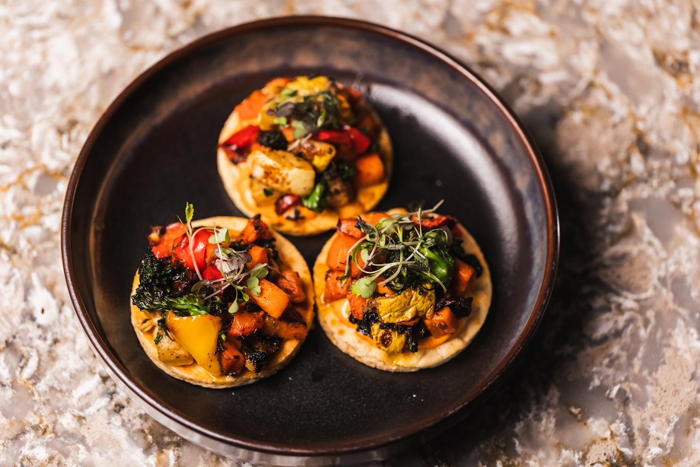 Three small, round flatbreads topped with colorful roasted vegetables and microgreens are arranged on a dark plate placed on a marbled surface.