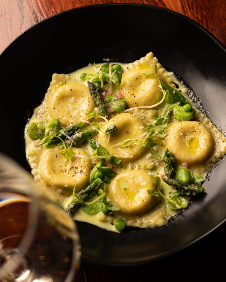 A black plate of round ravioli in a creamy sauce, garnished with green peas, asparagus, microgreens, and cracked pepper, with a blurred glass of wine in the foreground.