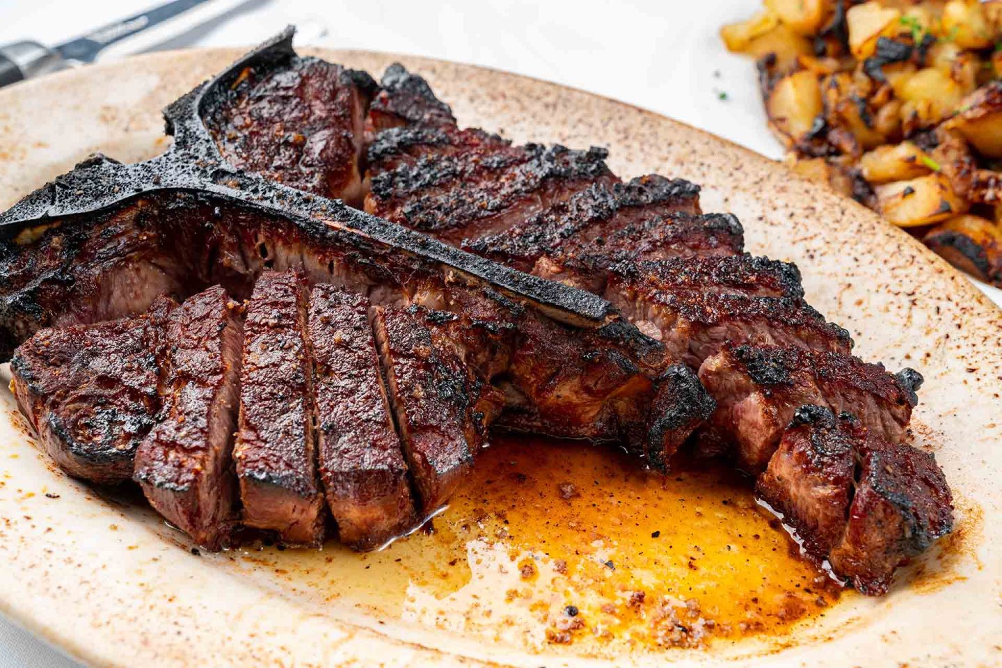 A sliced, juicy, grilled T-bone steak on a speckled plate with juices pooling around it, served alongside a portion of roasted potatoes.
