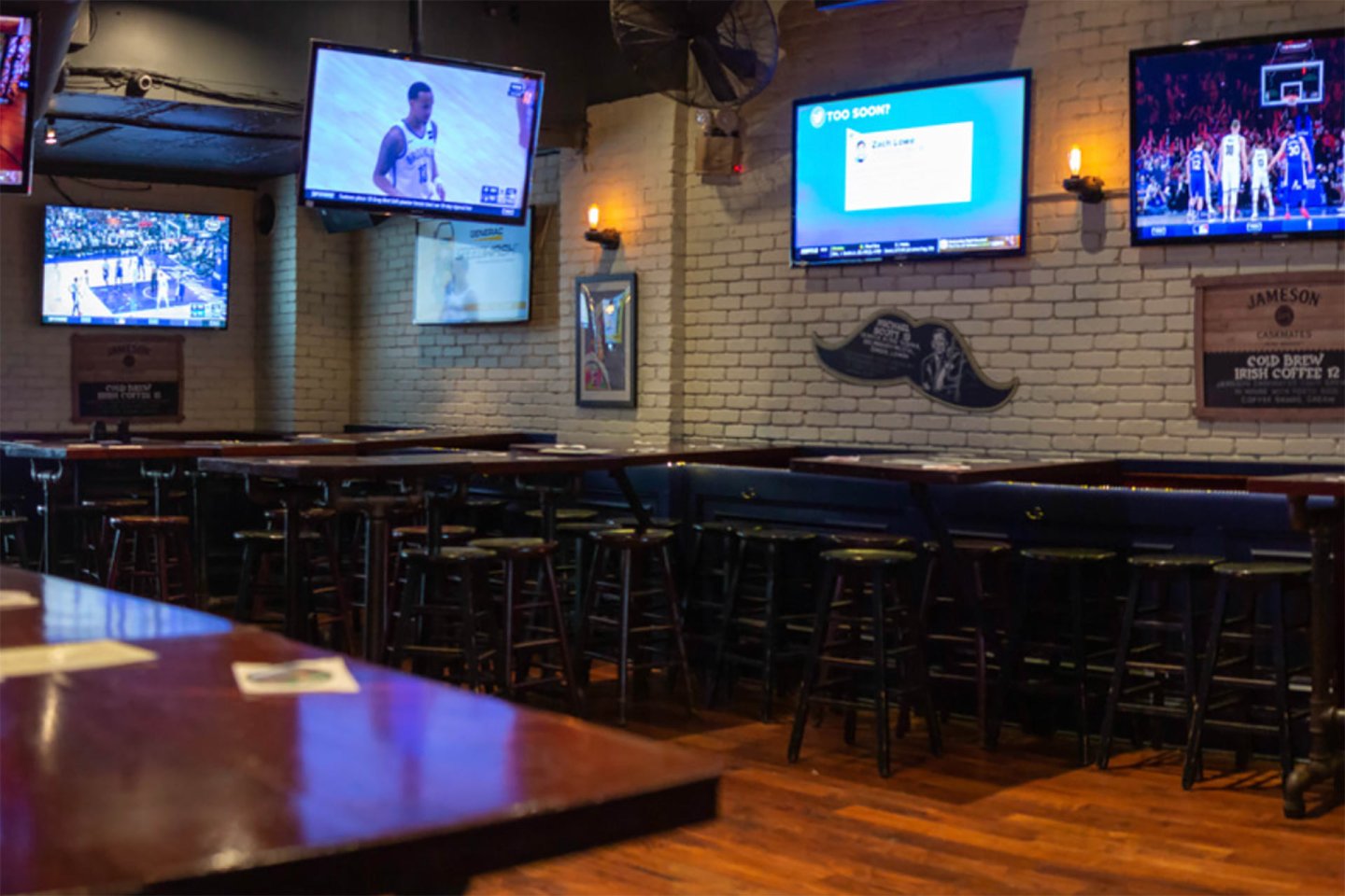 A dimly lit sports bar with several flat-screen TVs showing basketball games, high tables and stools, wooden floors, and a white brick wall with some framed decorations. The bar appears empty.