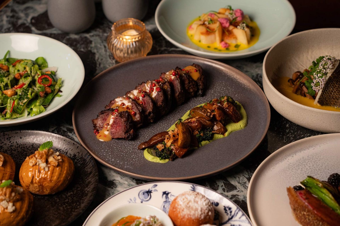 A table set with various gourmet dishes, including sliced steak with sauce and mushrooms, salad, fish, pastries, and artfully plated vegetables, all presented on elegant plates.