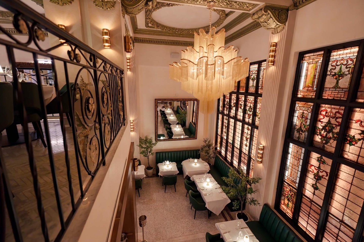 Elegant restaurant interior with high ceilings, large stained glass windows, green upholstered seating, white tablecloths, a grand chandelier, and a decorative mirror reflecting set tables below a balcony railing.