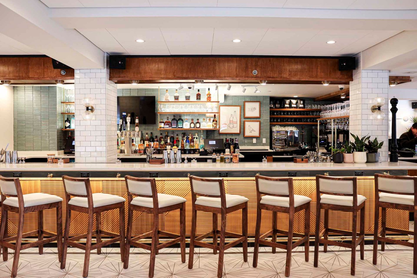 A modern bar with a row of seven wooden barstools with white cushions, a lighted counter, shelves stocked with bottles, glasses hanging above, white tile walls, and framed art behind the bar.