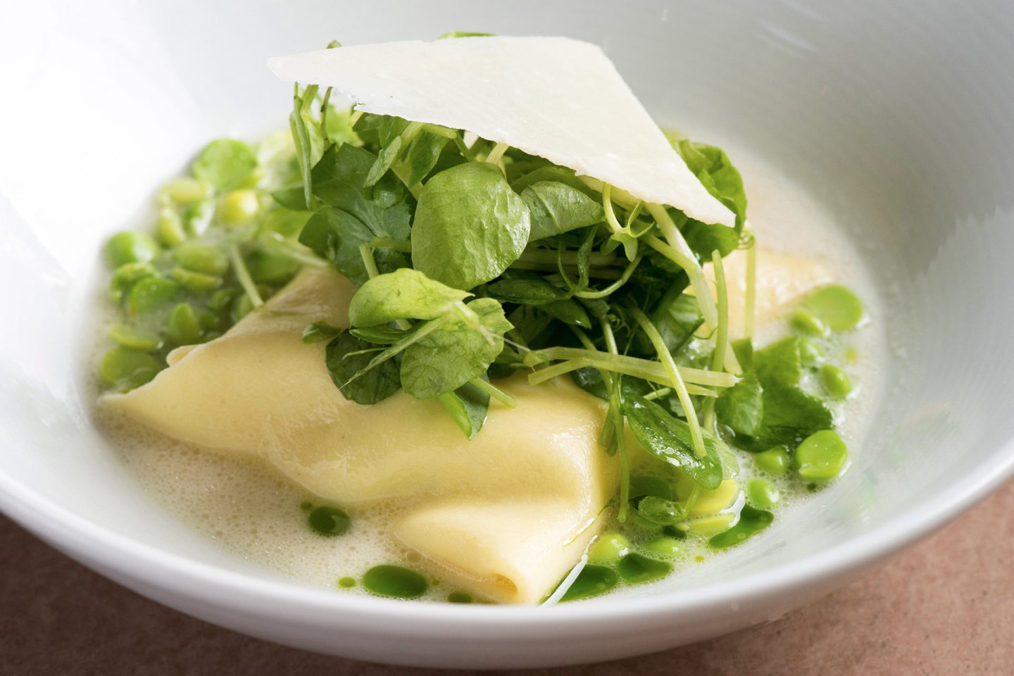 A white bowl containing ravioli topped with fresh green peas, pea shoots, and a thin slice of cheese, all sitting in a light green broth with small droplets of oil.