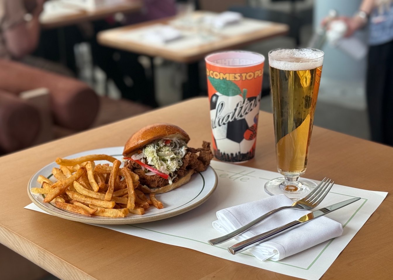 A spicy chicken sandwich with fries on a plate with a limited edition cup and draft beer
