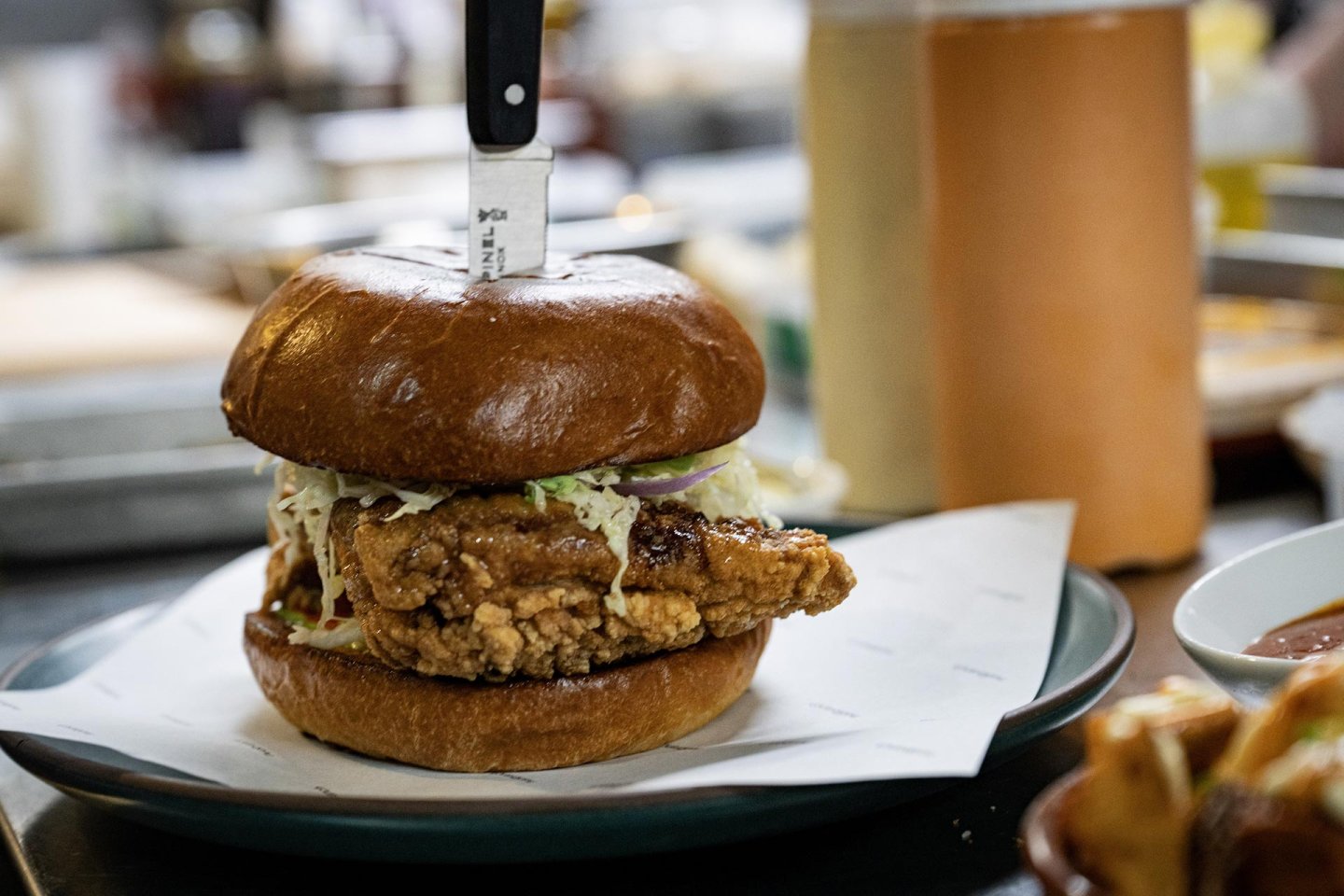 A fried chicken sandwich with coleslaw on a brioche bun sits on a plate, with a knife stabbed through the top. Blurred kitchen items and a bottle of sauce are visible in the background.