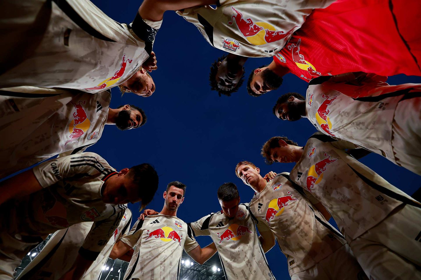 A group of soccer players in light uniforms with a Red Bull logo huddle together in a circle, looking down towards the camera against a dark evening sky.