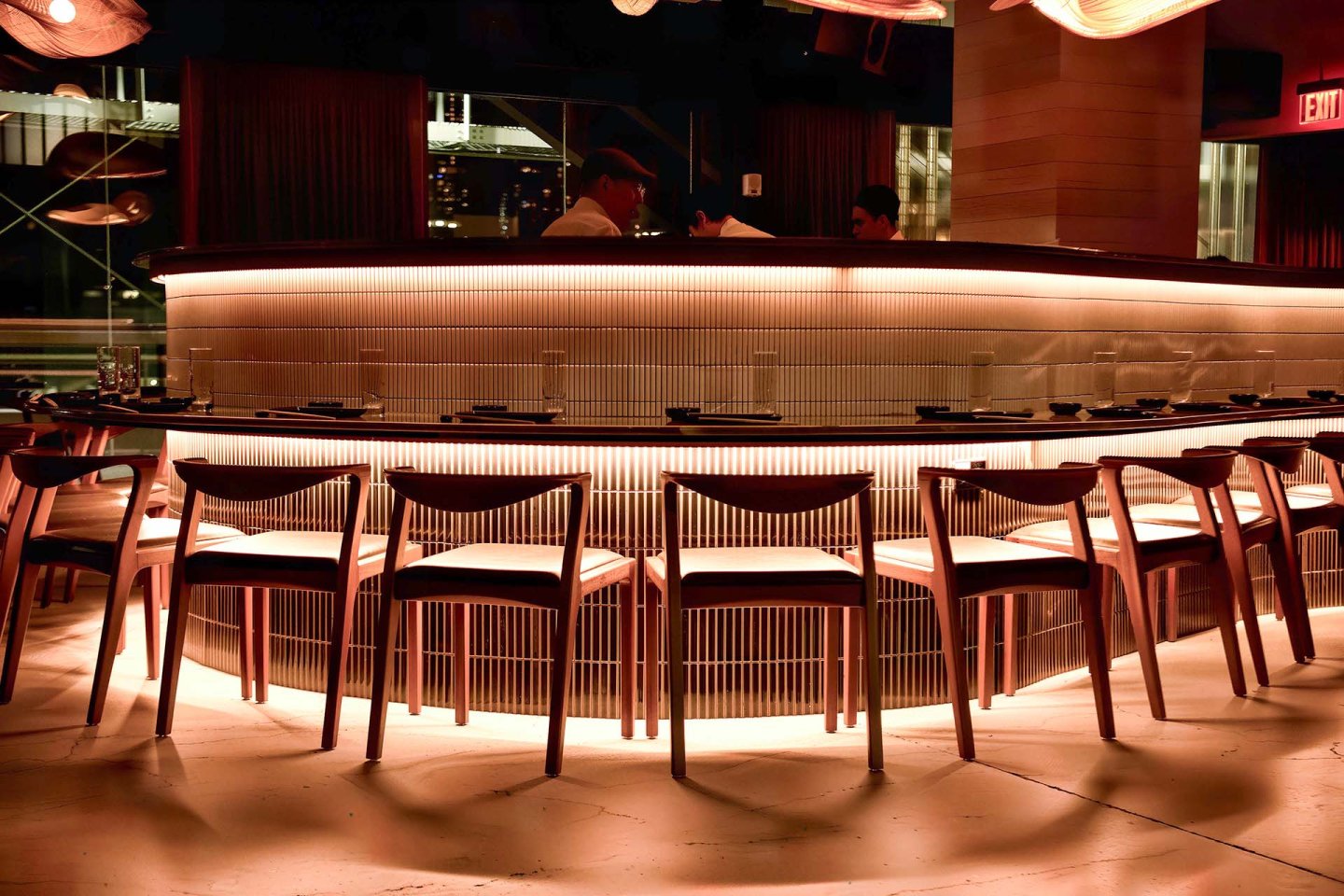 A modern, warmly lit bar with empty wooden chairs arranged in a semicircle, glowing under-counter lights, and two people standing behind the bar counter.