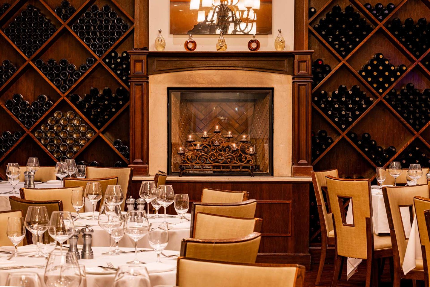Elegant restaurant with neatly set tables, wine glasses, and tan chairs. A decorative fireplace is centered on the back wall, flanked by shelves filled with wine bottles arranged in a diamond pattern. Warm lighting creates a cozy atmosphere.