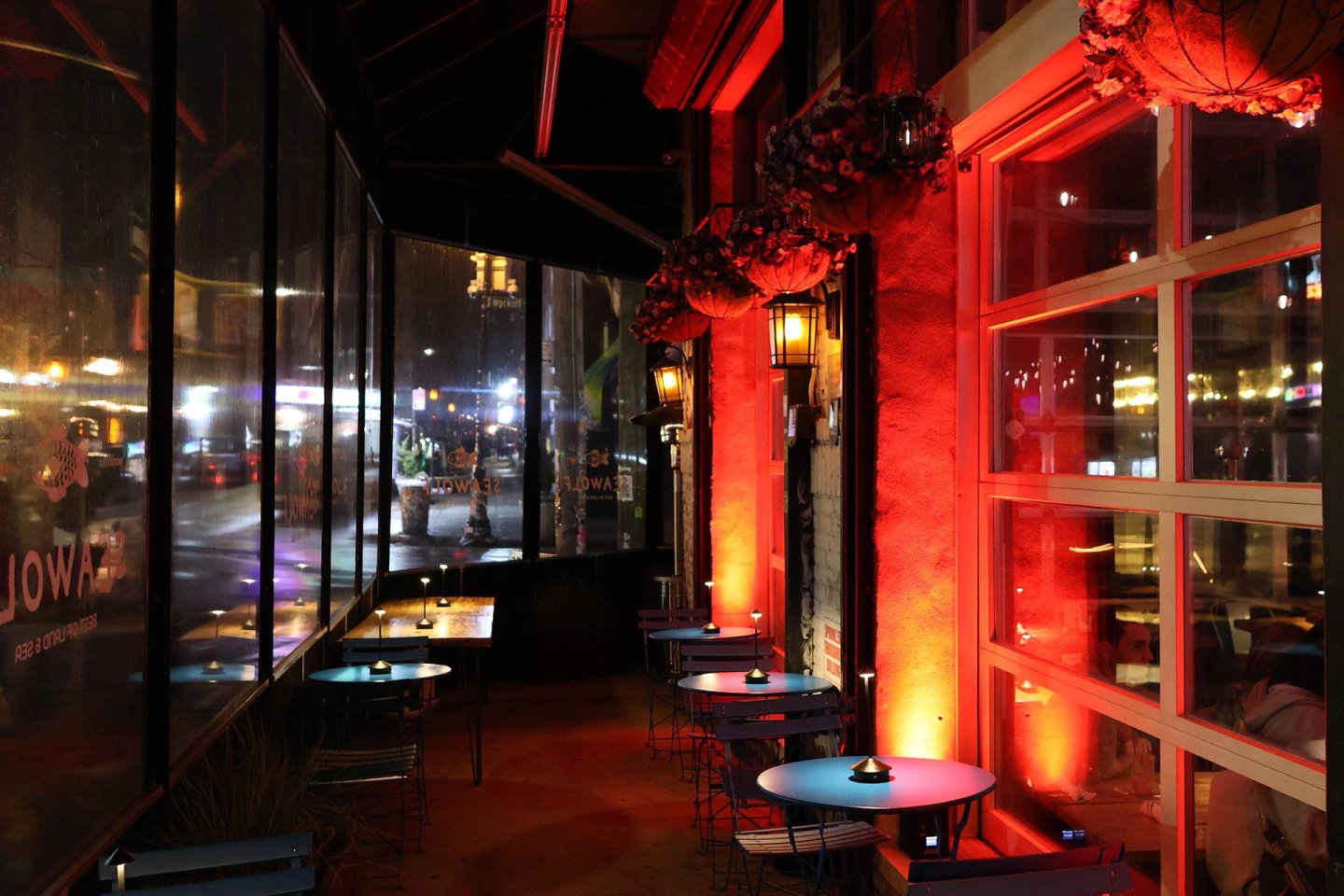 A cozy outdoor restaurant patio at night features round tables and chairs, lit by warm orange-red lights and lanterns. Reflections and city lights are visible through transparent plastic curtains on one side.