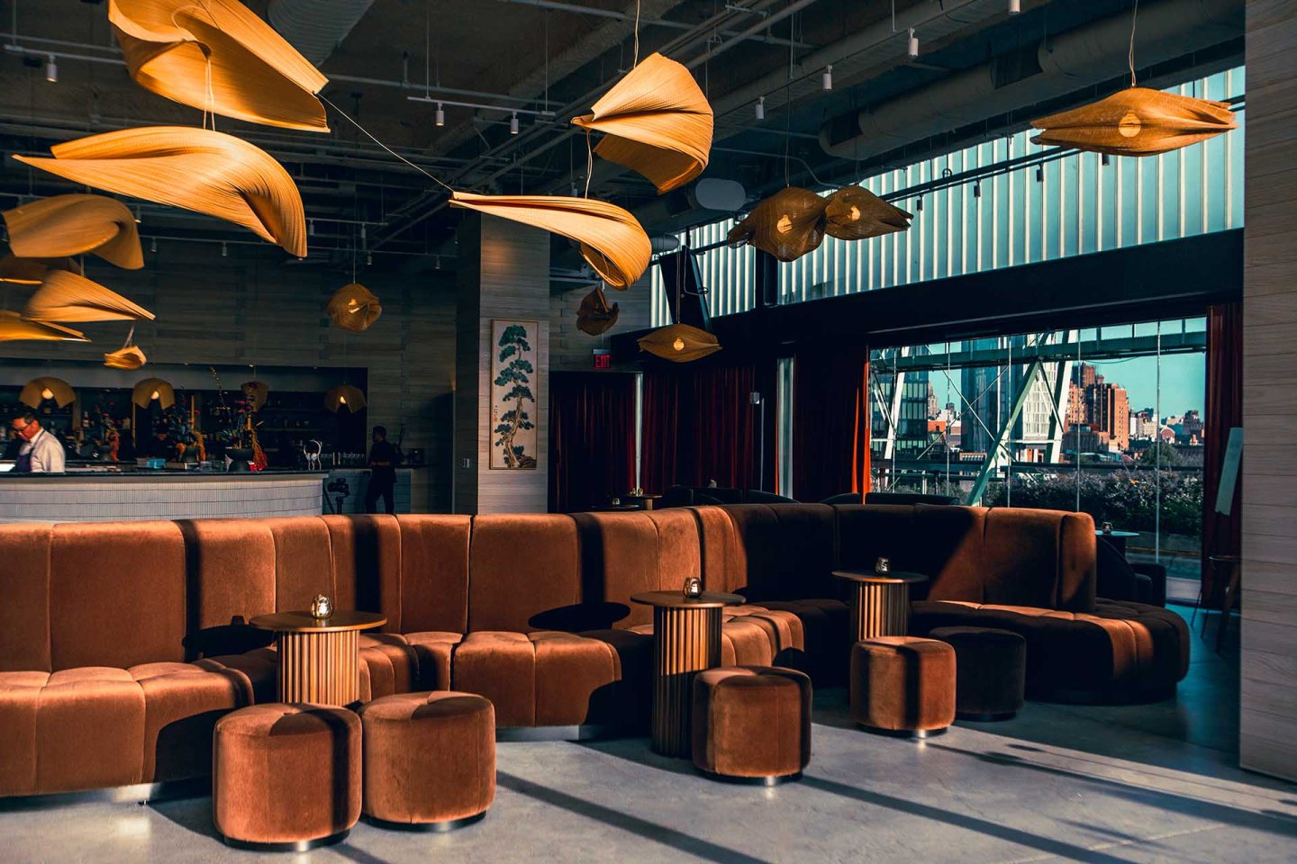 Modern lounge with brown velvet seating, small round tables, unique leaf-shaped ceiling lights, large windows, and a view of a cityscape in the background. Warm sunlight casts shadows across the stylish interior.