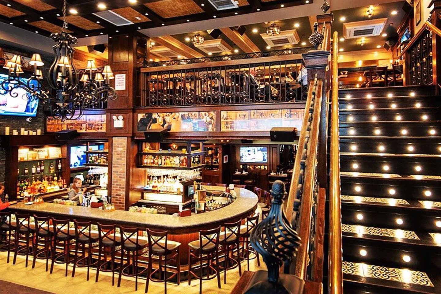 A spacious, warmly lit bar with a curved counter, high chairs, decorative chandeliers, and a sweeping staircase with illuminated steps leading to an upper mezzanine with more seating.