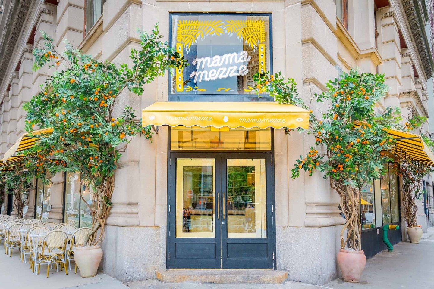 A corner restaurant with yellow-striped awnings, orange trees flanking the entrance, and a sign above the door that reads "mama mezze." Outdoor cafe seating with wicker chairs lines the sidewalk.