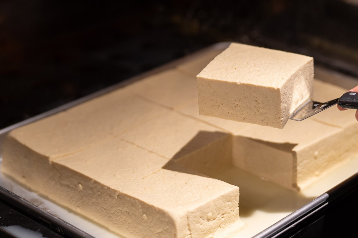A hand lifts a rectangular block of tofu from a tray filled with several large tofu blocks, some slightly submerged in liquid.