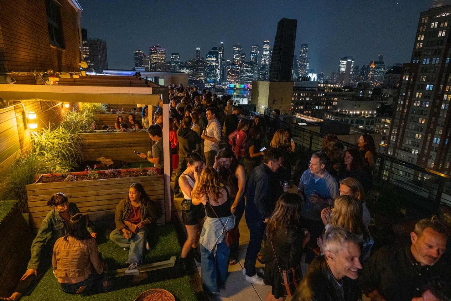 A crowded rooftop bar at night with people socializing, some sitting in booths and others standing. City skyscrapers are visible in the background, illuminated against the night sky.