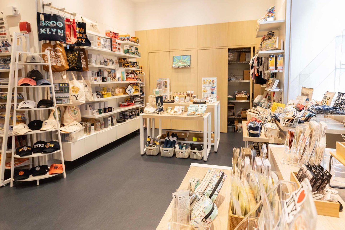 A brightly lit gift shop displays hats, bags, mugs, stationery, and souvenirs on shelves and tables. Items are neatly organized throughout the room, creating a welcoming and colorful shopping environment.