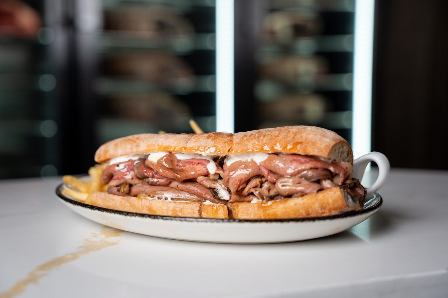 A large sandwich filled with sliced roast beef and melted cheese is served on a white plate with a side of fries, set on a white countertop.