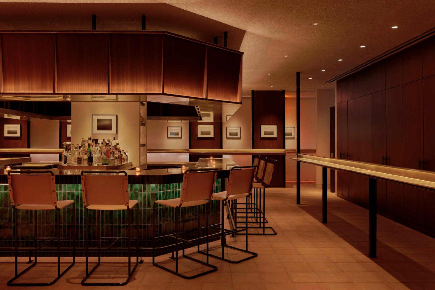 A modern bar with sleek wooden finishes, featuring a curved counter with high chairs. Soft ambient lighting illuminates the minimalist decor, including framed art on the walls. The space has a warm and inviting atmosphere.