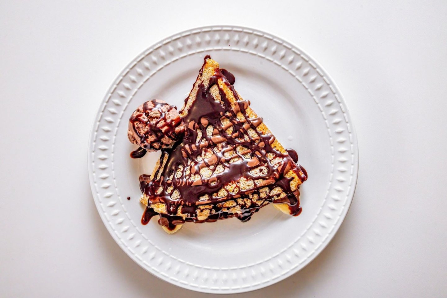 A triangular waffle topped with chocolate sauce and a scoop of chocolate ice cream sits on a white plate against a white background.