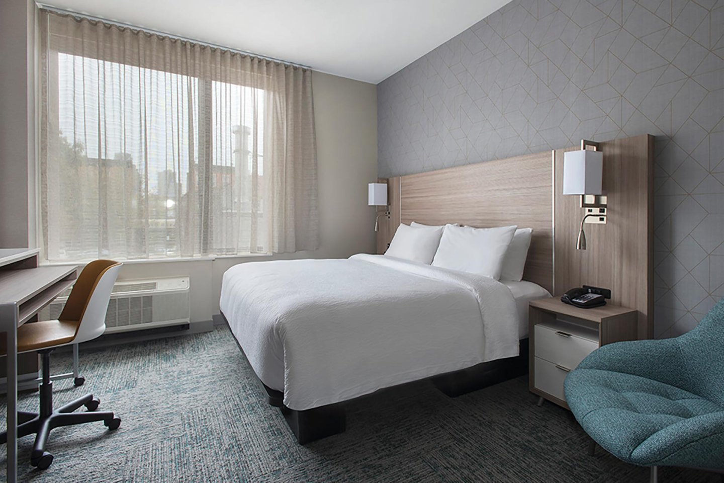 A modern hotel room with a large bed, white bedding, light wood headboard, two nightstands with lamps, a blue armchair, a desk with chairs, and a large window with sheer curtains letting in natural light.