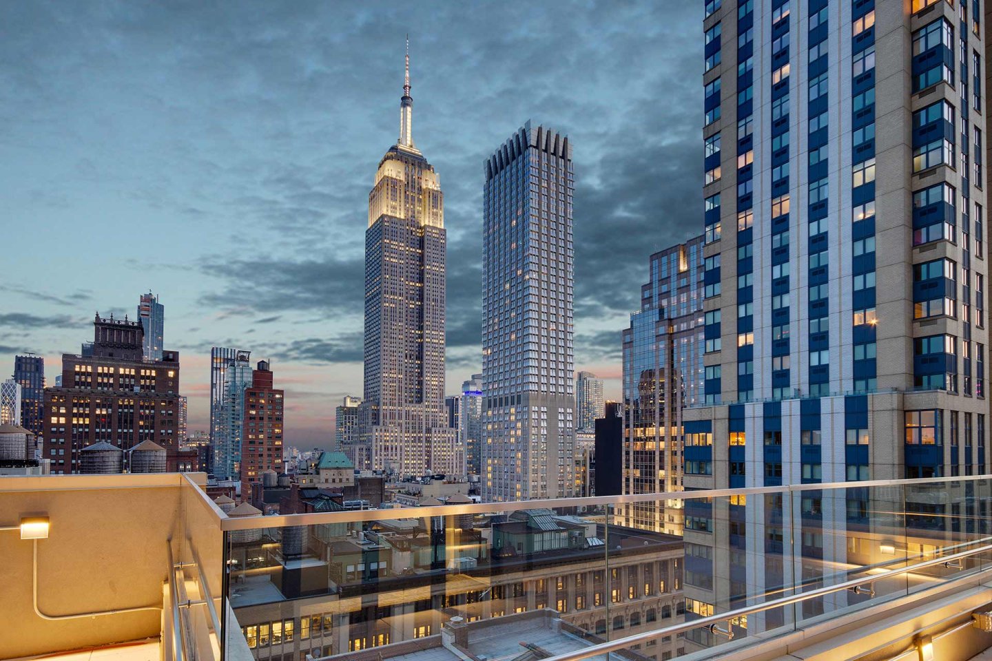 Rooftop view of midtown Manhattan at dusk, featuring the Empire State Building and surrounding skyscrapers under a partly cloudy sky, with glass railings in the foreground.