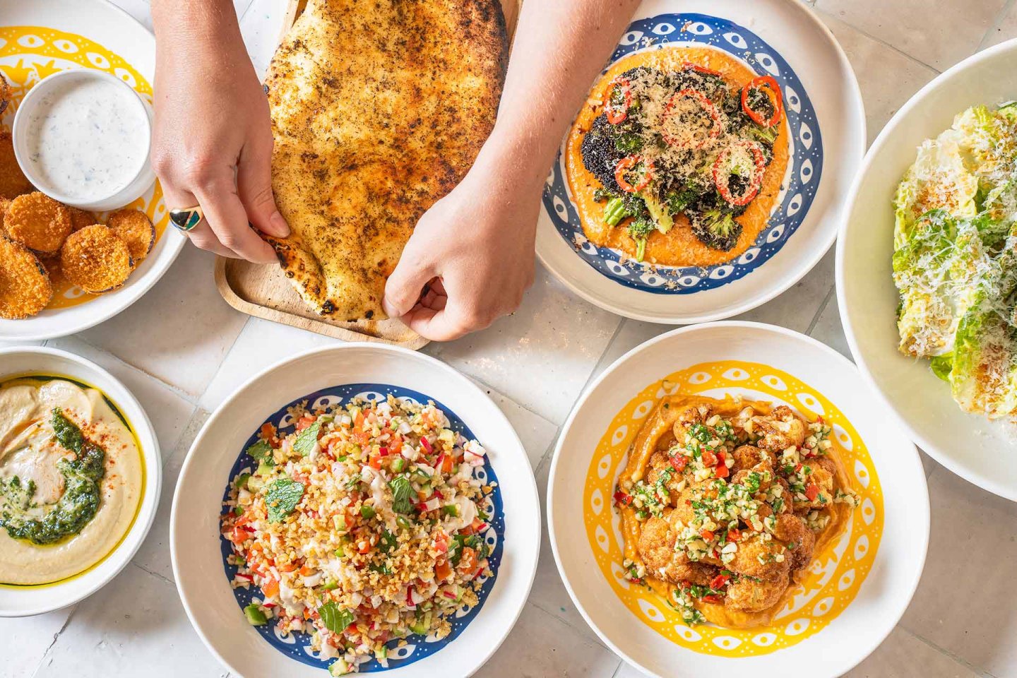 A table with colorful dishes including flatbread, salad, hummus, falafel, curry, and a creamy dip. Two hands are pulling apart the flatbread over the spread of vibrant Middle Eastern or Mediterranean food.