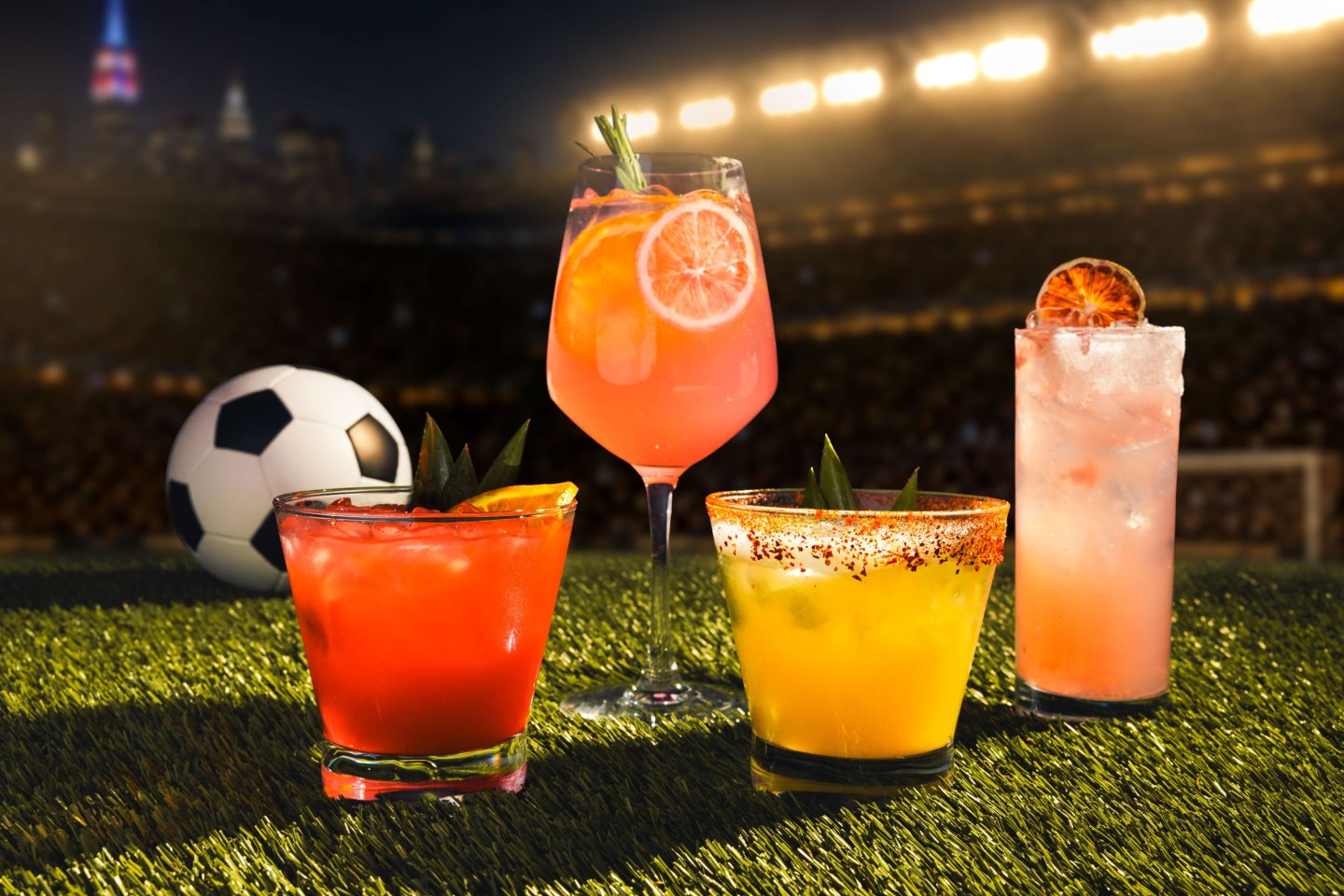 Four colorful specialty cocktails displayed on artificial turf in front of a stadium backdrop, with a soccer ball and the New York City skyline visible in the background. The drinks feature vibrant red, pink, yellow, and blush-colored presentations with citrus garnishes, reflecting a soccer-inspired cocktail lineup.