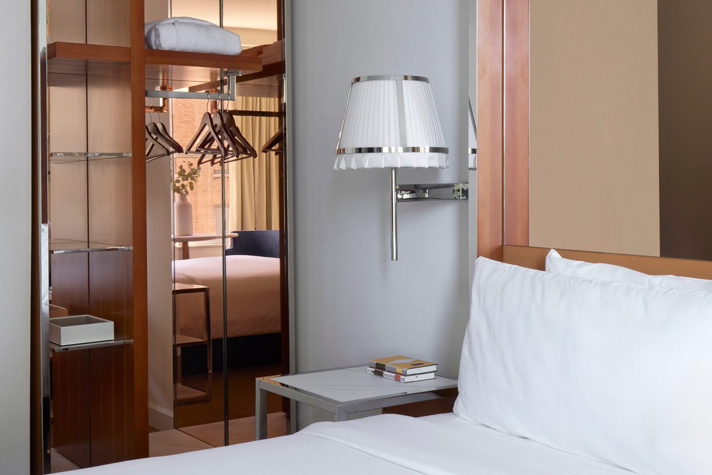A modern hotel room with a neatly made bed, white linens, a wall-mounted lamp, a small side table with books, an open wardrobe with hangers, and a view into another bedroom with a window and flowers.