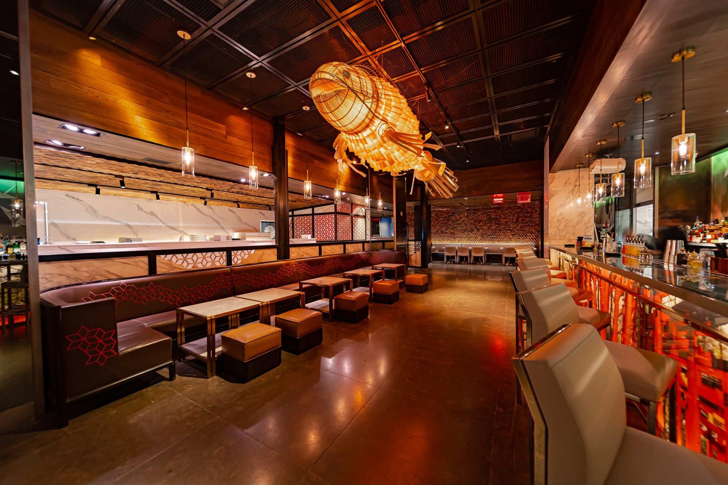 Modern restaurant interior with a large, illuminated fish-shaped light fixture, cushioned seating, a long bar with stools, and warm ambient lighting. The space features wood accents and a contemporary design.
