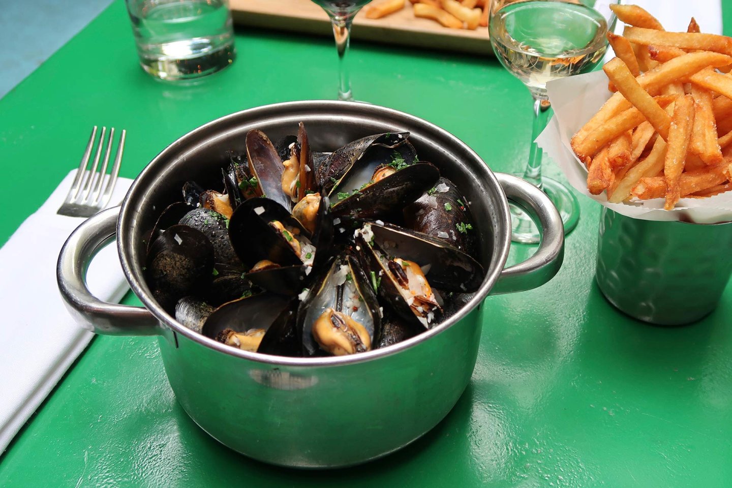 A pot of steamed mussels with herbs sits on a green table next to a cup of golden French fries, a glass of white wine, and a fork and napkin.
