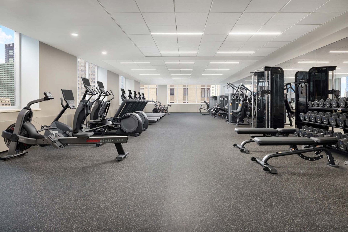 A spacious, modern gym with cardio machines like treadmills and ellipticals on the left, weightlifting equipment and racks of dumbbells on the right, and large windows letting in natural light.