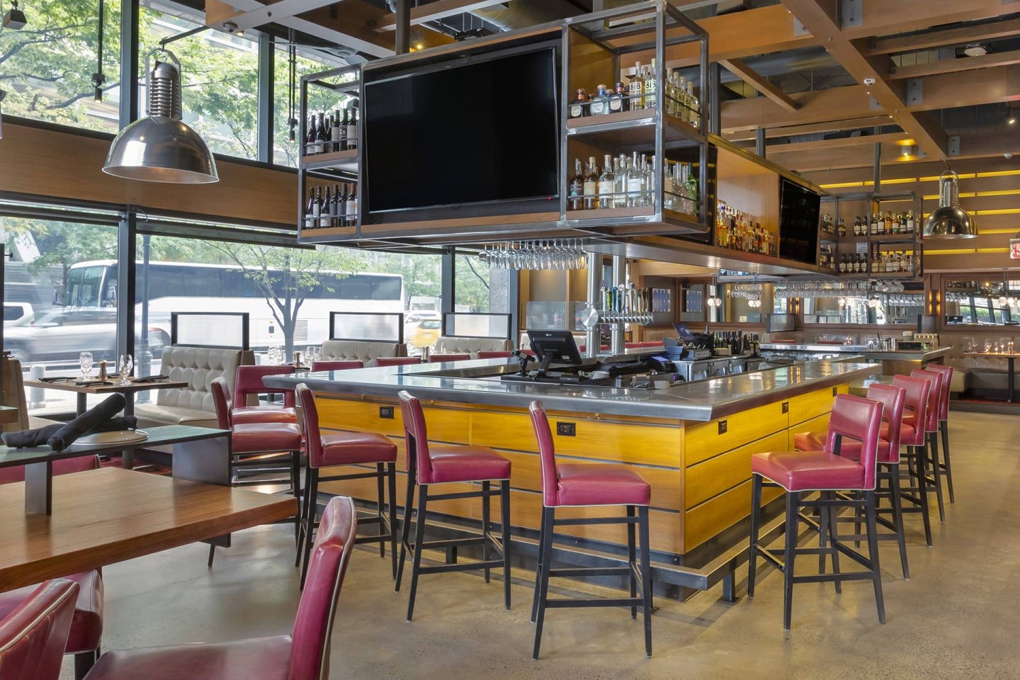 A modern restaurant bar with high red chairs, a large TV above the counter, bottles of liquor on shelves, and large windows letting in natural light. Tables and booths are visible in the background.