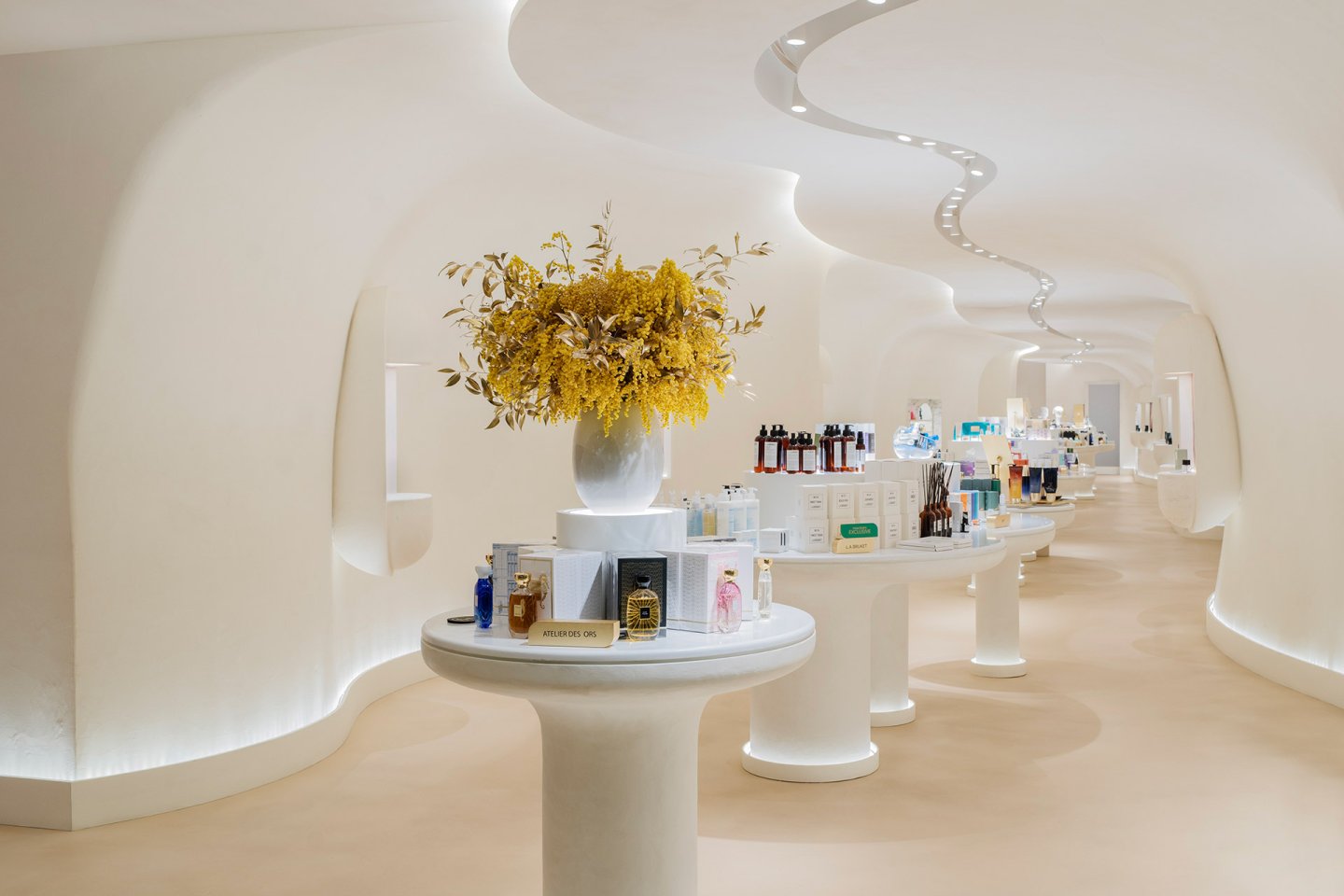A modern, elegant store with white curved walls and ceiling, featuring sleek display tables holding various beauty products and a vase of yellow flowers in the foreground. Soft lighting creates a calm, luxurious atmosphere.