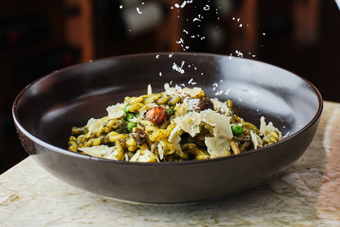 A dark bowl filled with pasta topped with pesto, vegetables, mushrooms, and shaved parmesan, with cheese being freshly grated and falling onto the dish. The bowl sits on a marble surface.